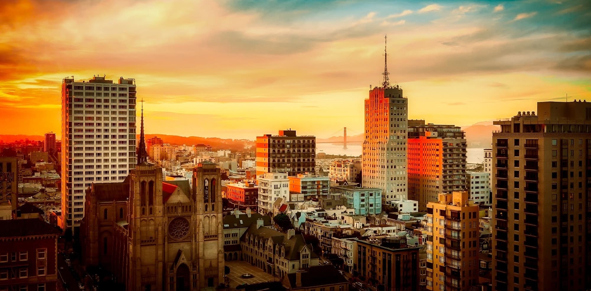 Sunset view of a city skyline with historic and modern buildings, featuring a church with tall spires in the foreground and a bridge visible in the distance under a golden sky.