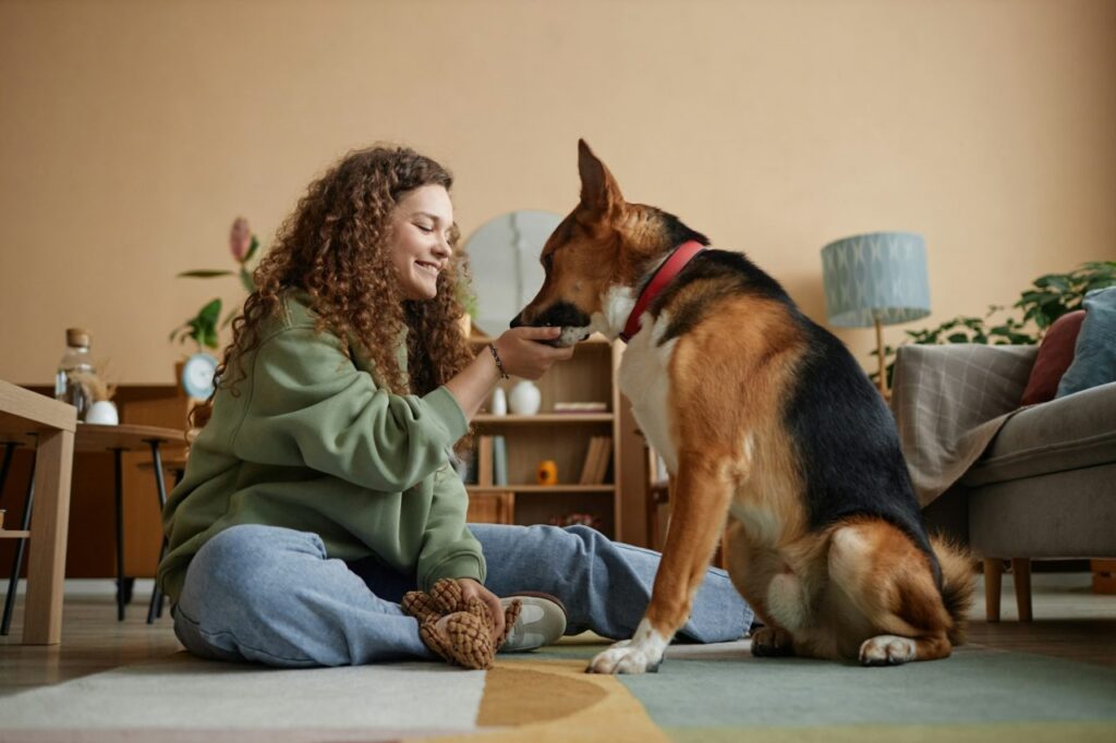 A smiling woman with curly hair sits on the floor indoors, wearing a green sweatshirt and jeans, feeding a large dog with a red collar from her hand, surrounded by cozy home furnishings including a lamp, plants, and a couch.