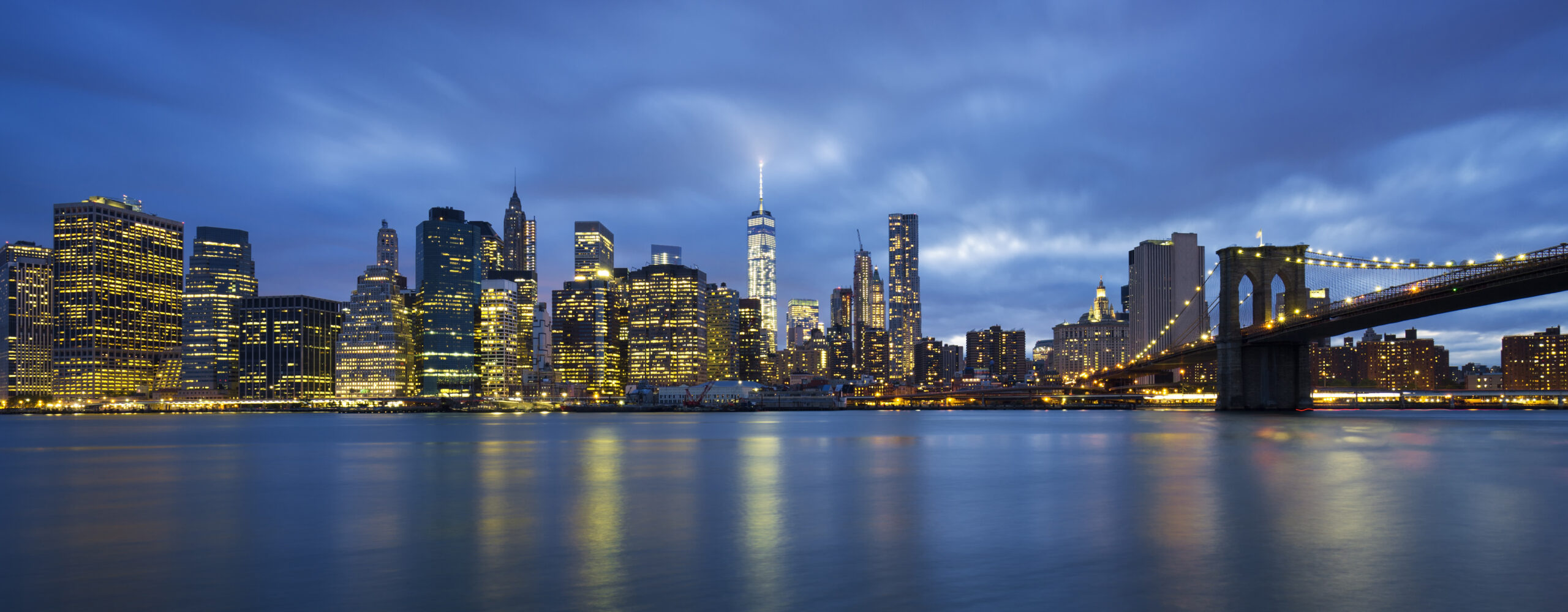 Nighttime panoramic view of Manhattan skyline with illuminated skyscrapers and the Brooklyn Bridge on the right, reflecting on the calm water under a cloudy blue sky.